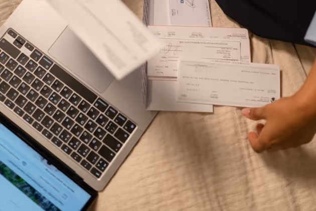 Person organizing multiple bills next to a laptop on a beige quilted surface.