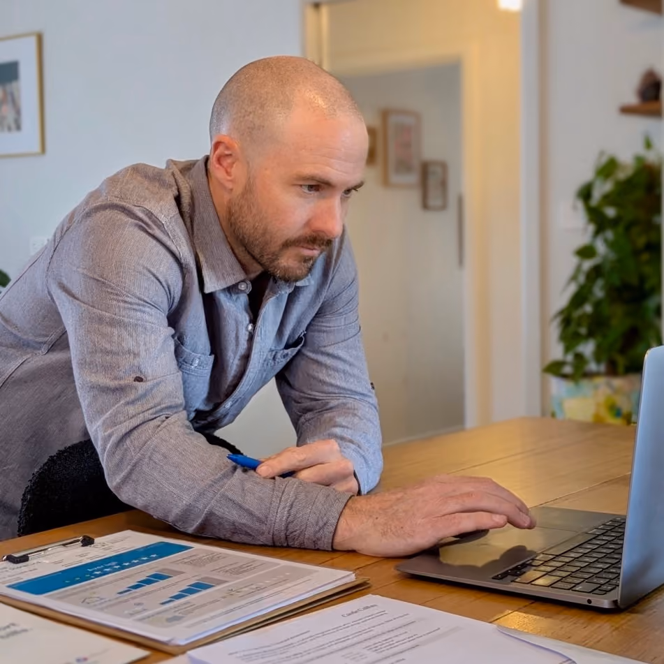 Man in gray shirt working on a laptop at a table with documents and a pen.